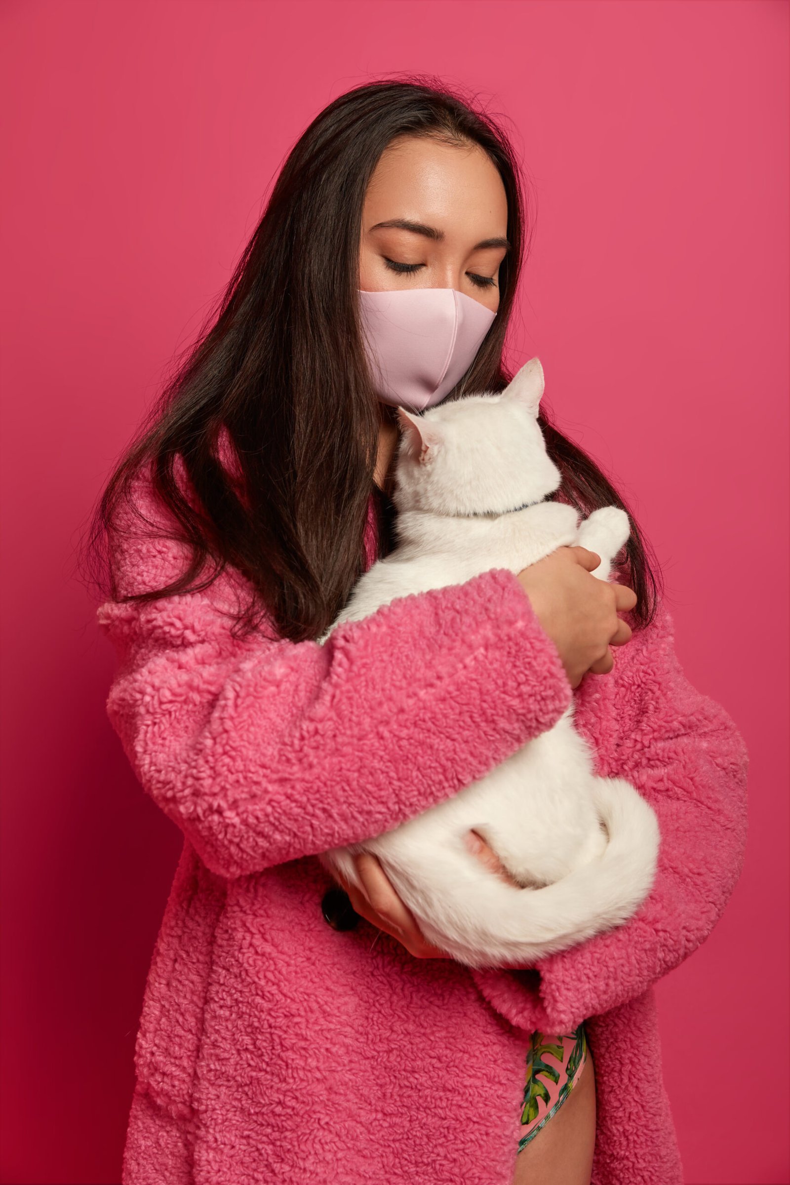 Photo of brunette woman wears protective facial mask at home, tries stop virus, cares about white cat, wears coat, isolated on pink studio background, being sick, has allergy on domestic animals